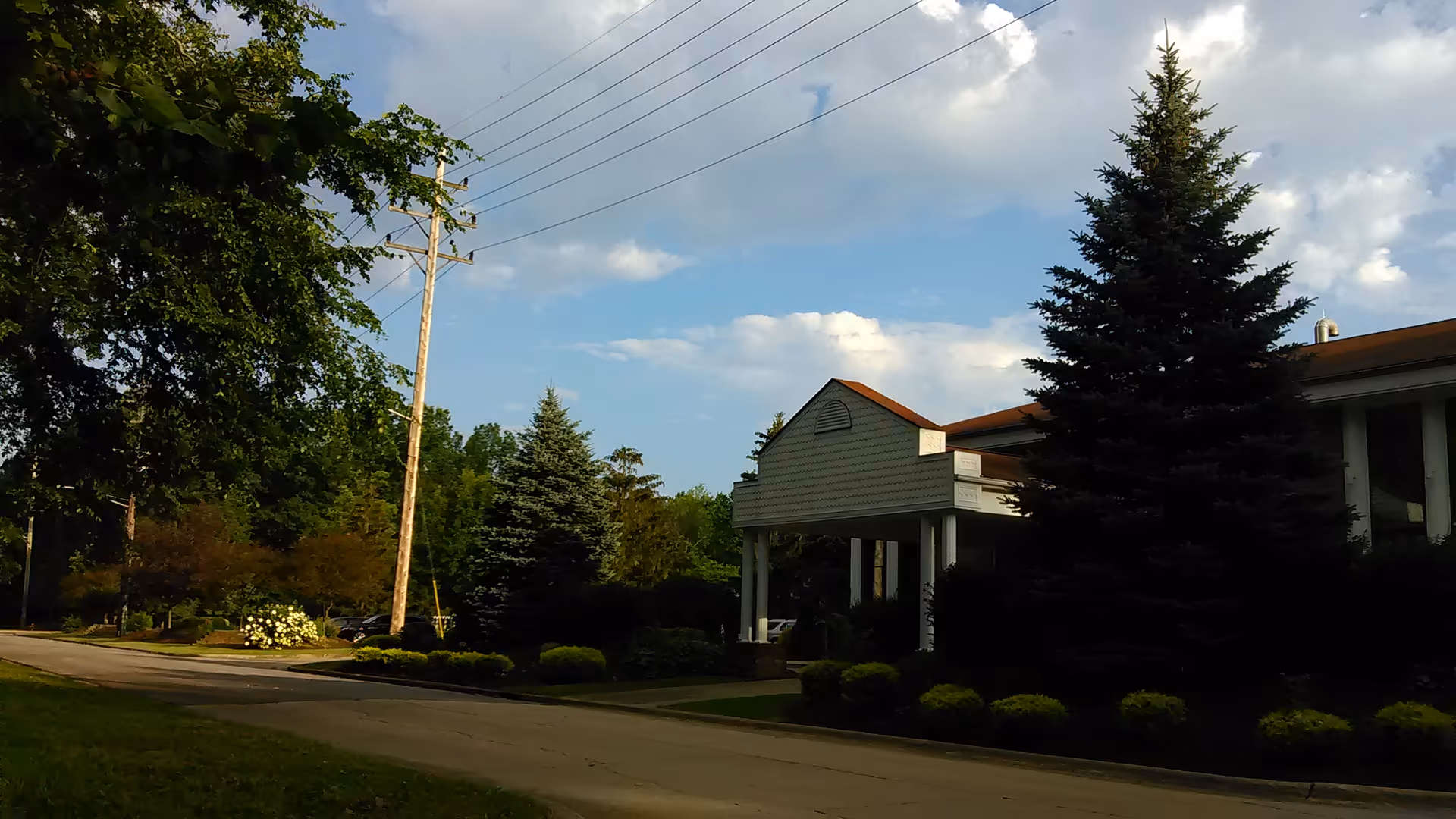 Exterior view of a facility with a covered entrance, trees, and manicured bushes beside a street.