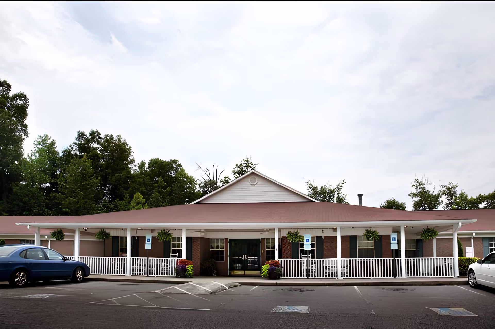 Front exterior of a single-story senior living building with a covered porch, white railings, hanging planters, and parked cars.