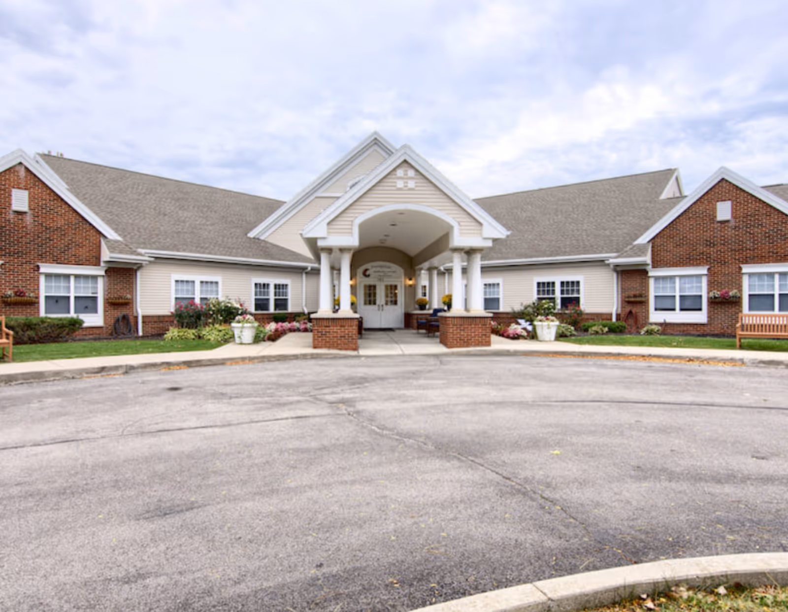 Front exterior view of Peregrine Senior Living at Orchard Park, a single-story building with a covered entrance supported by white columns, brick and beige siding, surrounded by landscaped greenery and flowers.