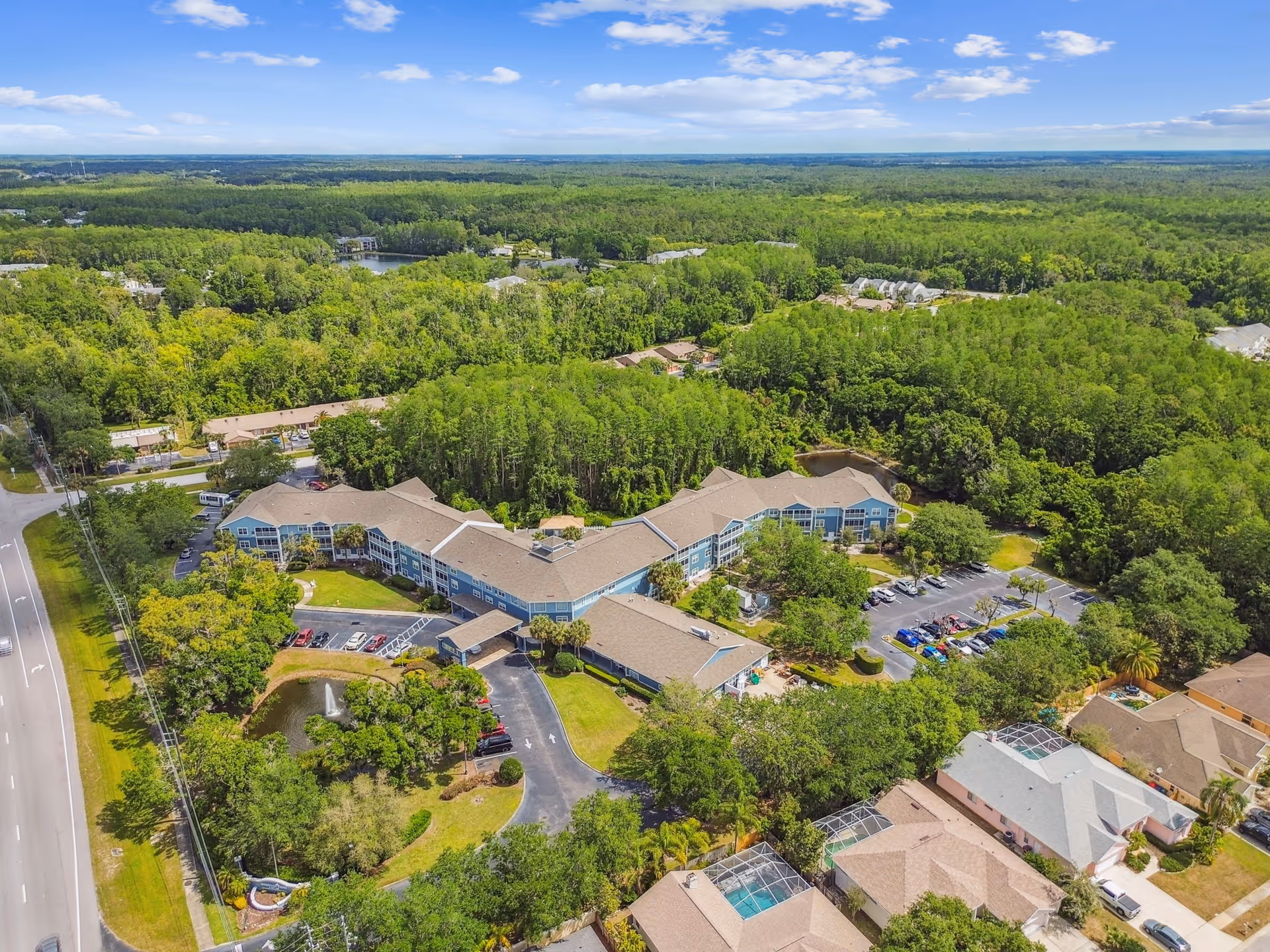 Aerial view of Addington Place of East Lake senior living facility surrounded by lush green trees and residential houses. The facility features a large, multi-wing building with parking lots, a small pond with a fountain, and well-maintained landscaping. A road runs along the left side of the property.