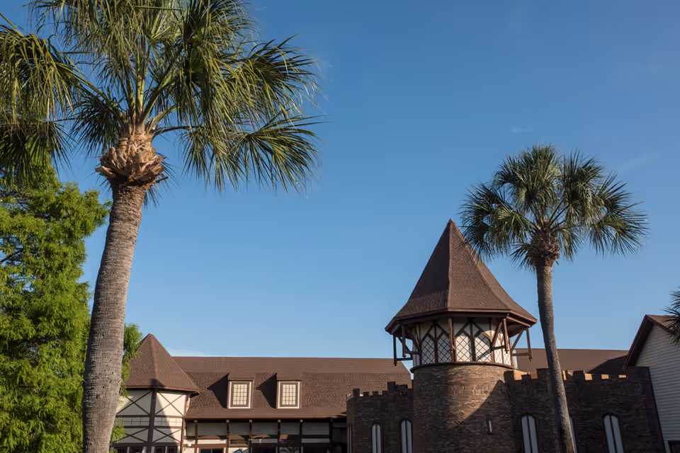 Exterior view of a castle-like building with a turret and palm trees under a clear blue sky.