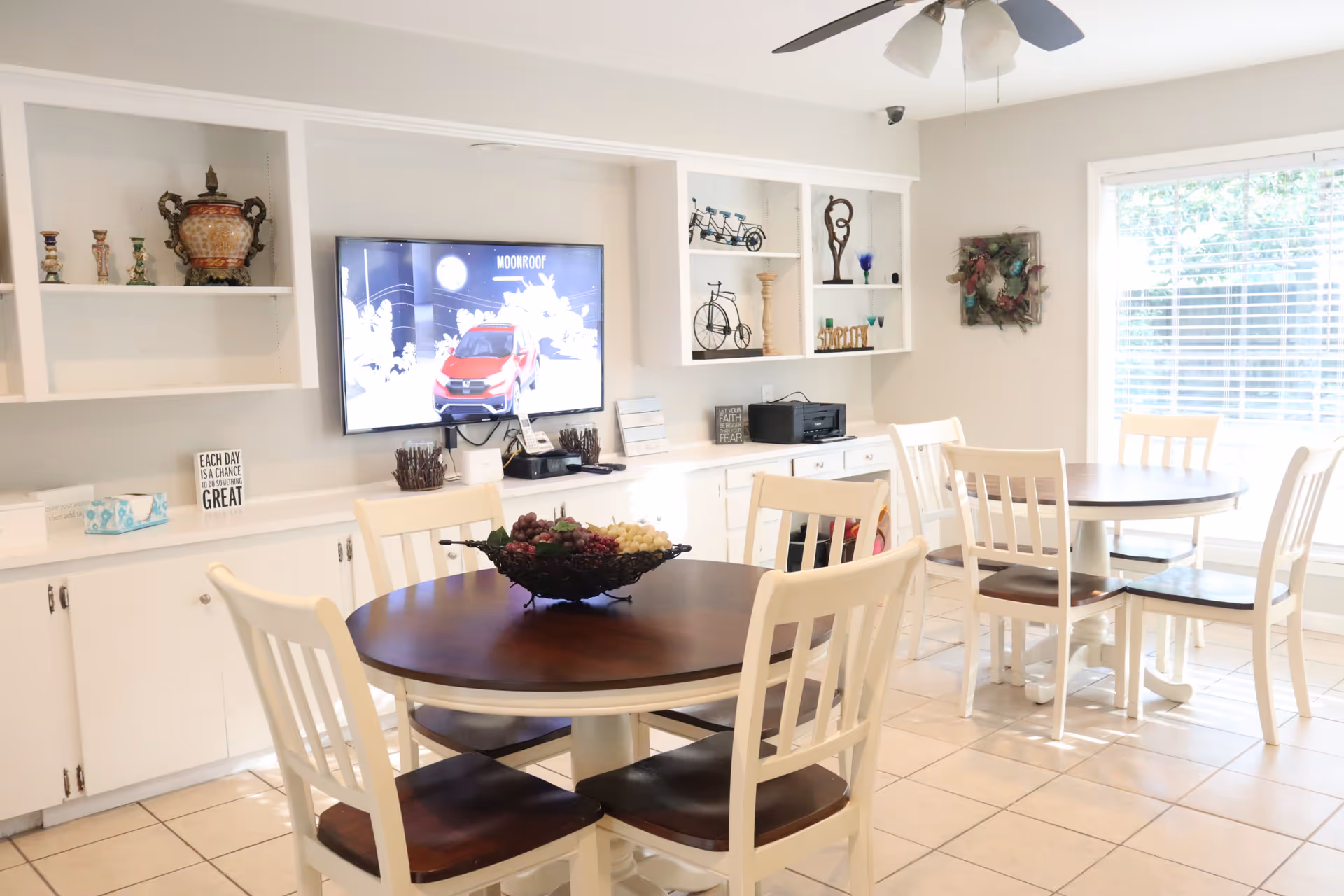 A bright dining area with two round wooden tables surrounded by white chairs with dark wooden seats. A decorative bowl with artificial grapes is placed on the nearest table. The room features built-in white shelves and cabinets along one wall, decorated with various ornaments and a mounted flat-screen TV displaying an image of a red car and the word 'MOONROOF'. A large window with white blinds allows natural light to fill the space.
