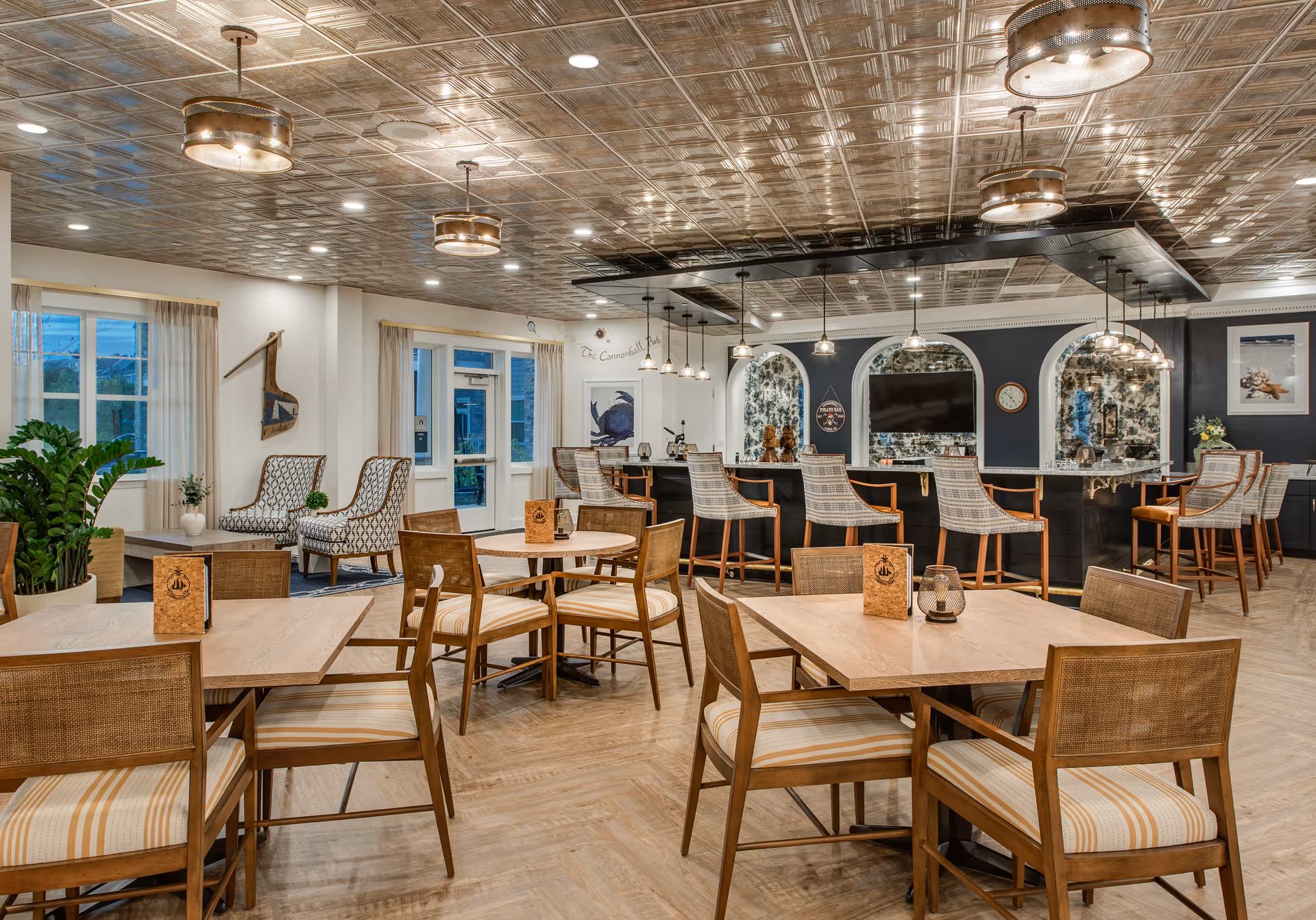 A spacious and well-lit dining area in a senior living facility featuring wooden tables and chairs with striped cushions. The room has a decorative tin ceiling with multiple pendant lights and ceiling fixtures. In the background, there is a bar area with high chairs, a large TV mounted on a dark accent wall with arched mirrors, and some decorative items. Large windows with light curtains allow natural light to enter the room.