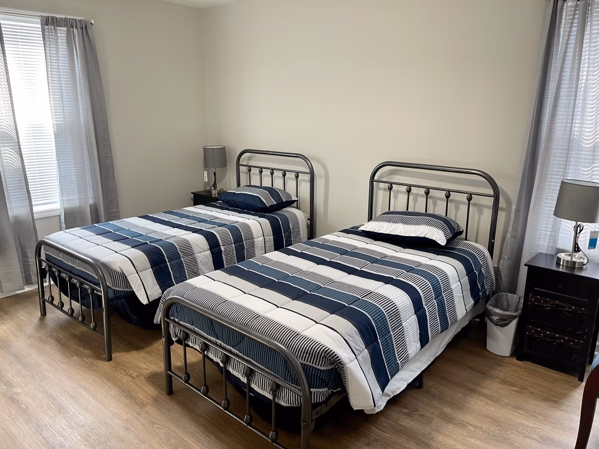 A bedroom with two single beds featuring metal frames and blue, white, and black striped bedding. Each bed has a pillow and is positioned next to a window with gray curtains. There are two black nightstands, each with a gray lamp, and a small trash bin next to one of the nightstands. The room has light-colored wooden flooring and plain light-colored walls.