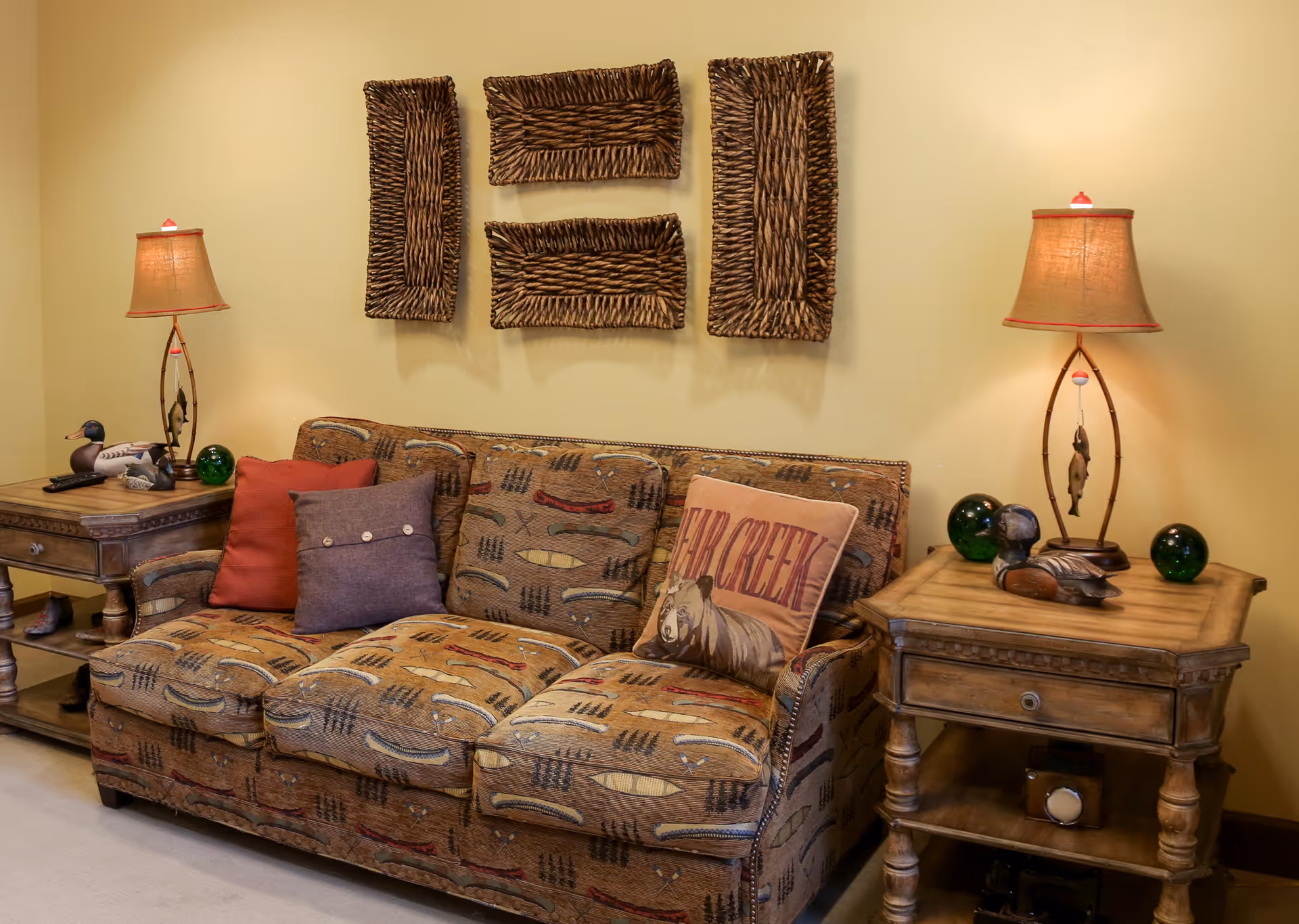 A cozy living room area with a patterned sofa adorned with three decorative pillows, two wooden side tables each holding a lamp with a beige shade, and decorative items including duck figurines and green glass spheres. Above the sofa, there are four rectangular woven wall hangings arranged in a rectangular pattern on a light yellow wall.