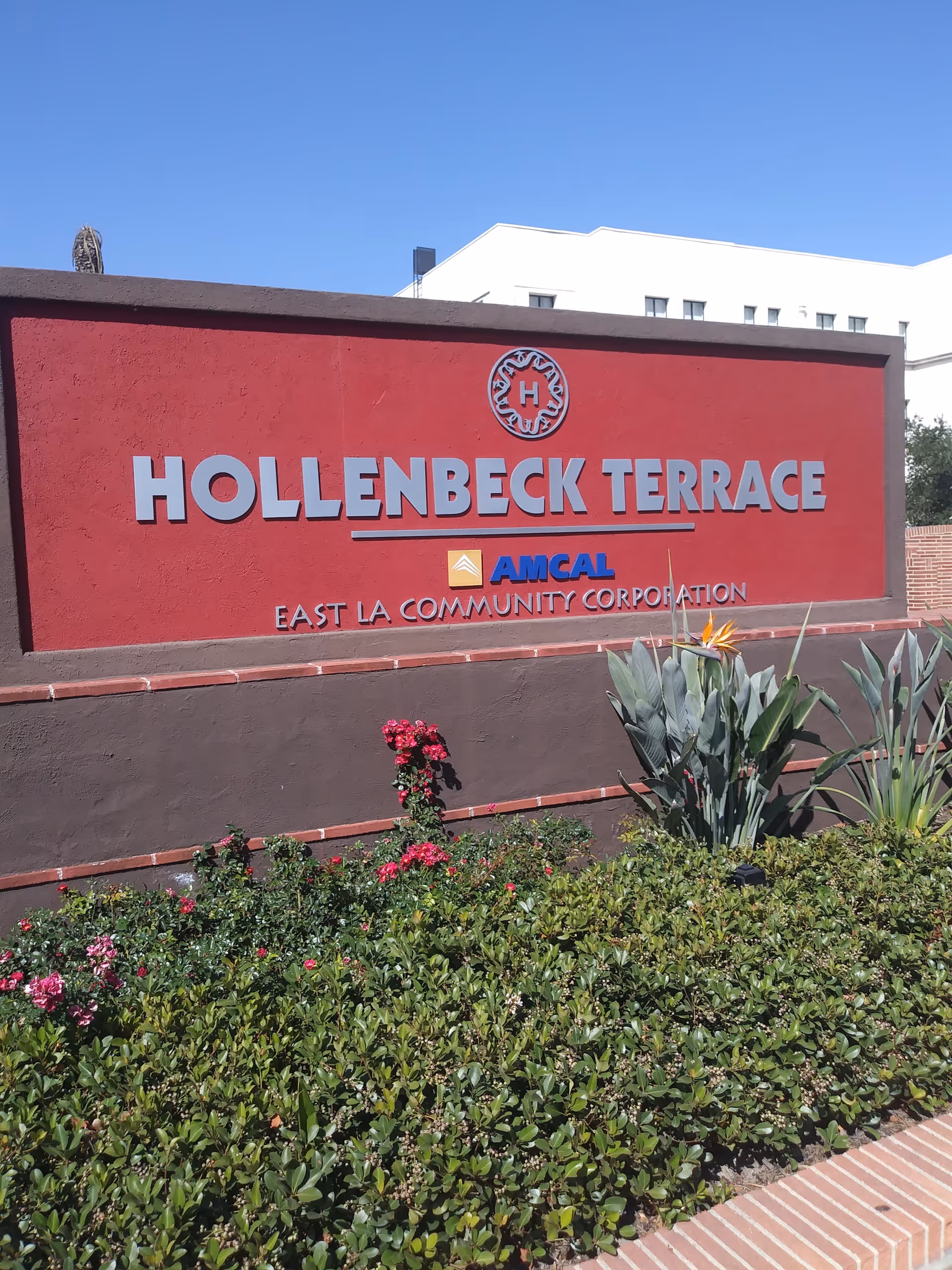 Red Hollenbeck Terrace sign with greenery and a white building behind.