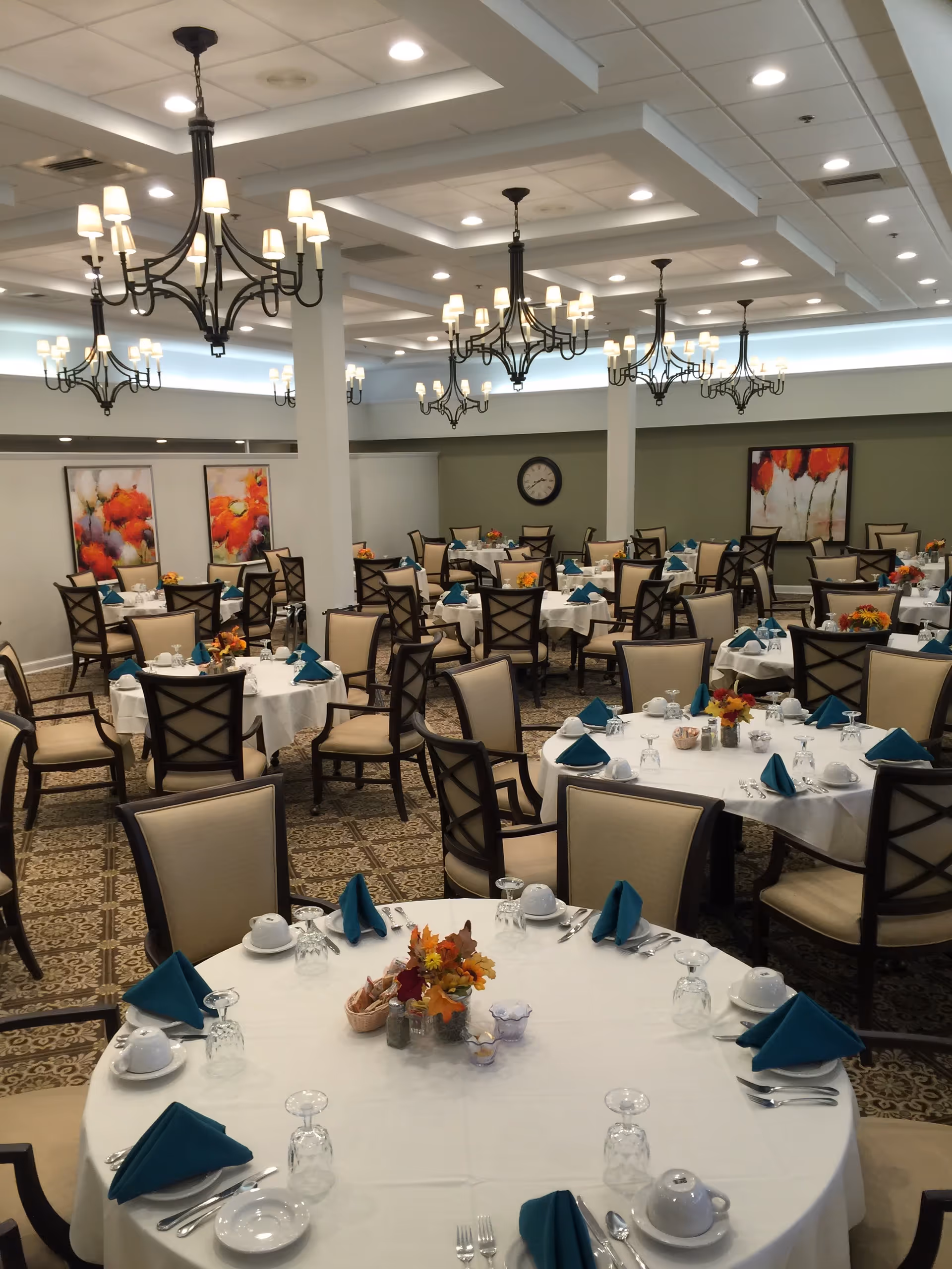 Spacious dining room with round tables set for meals, teal napkins, and elegant chandeliers.