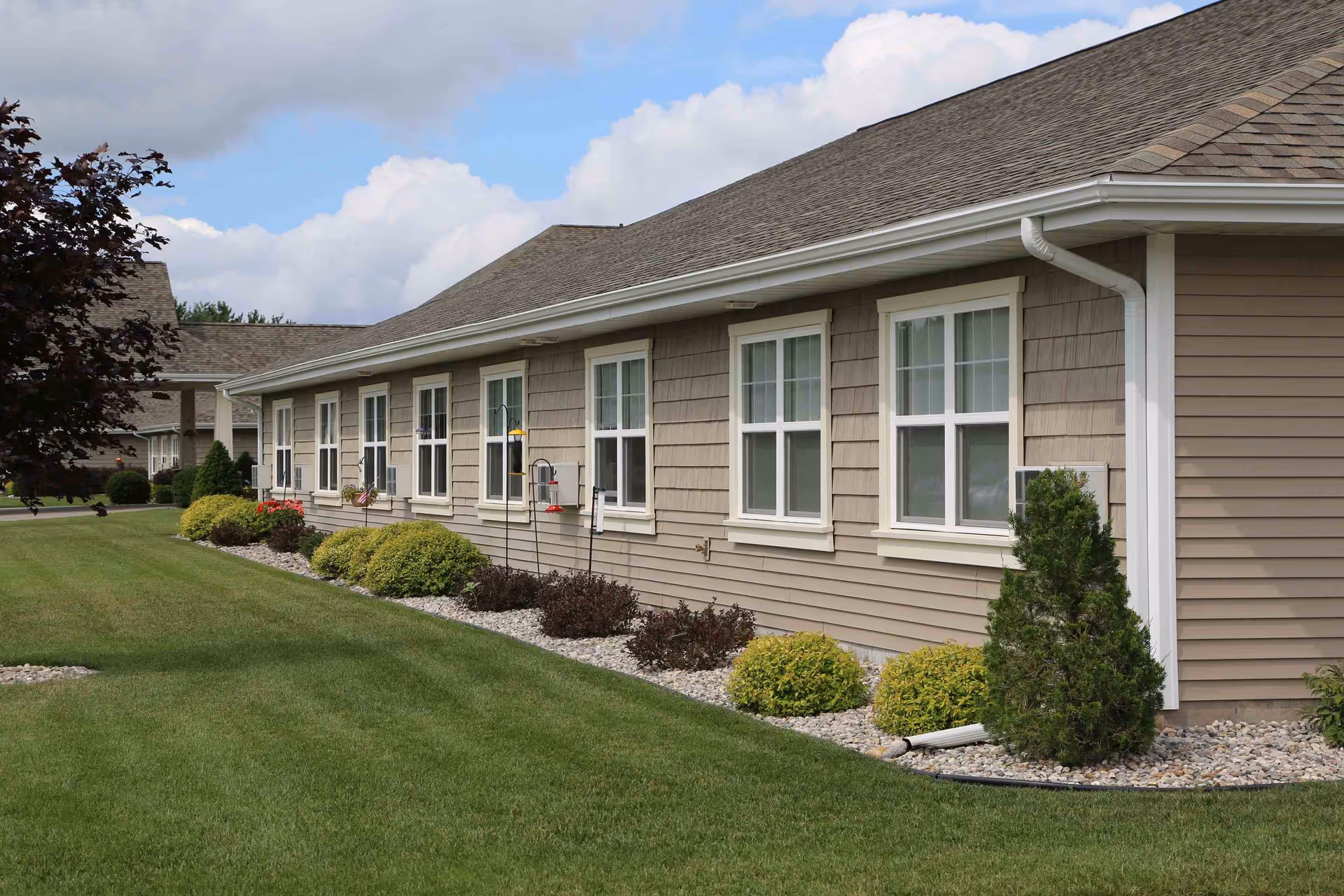 Side exterior of a single-story residential building with a row of windows, shrubs, and a manicured lawn under a partly cloudy sky.