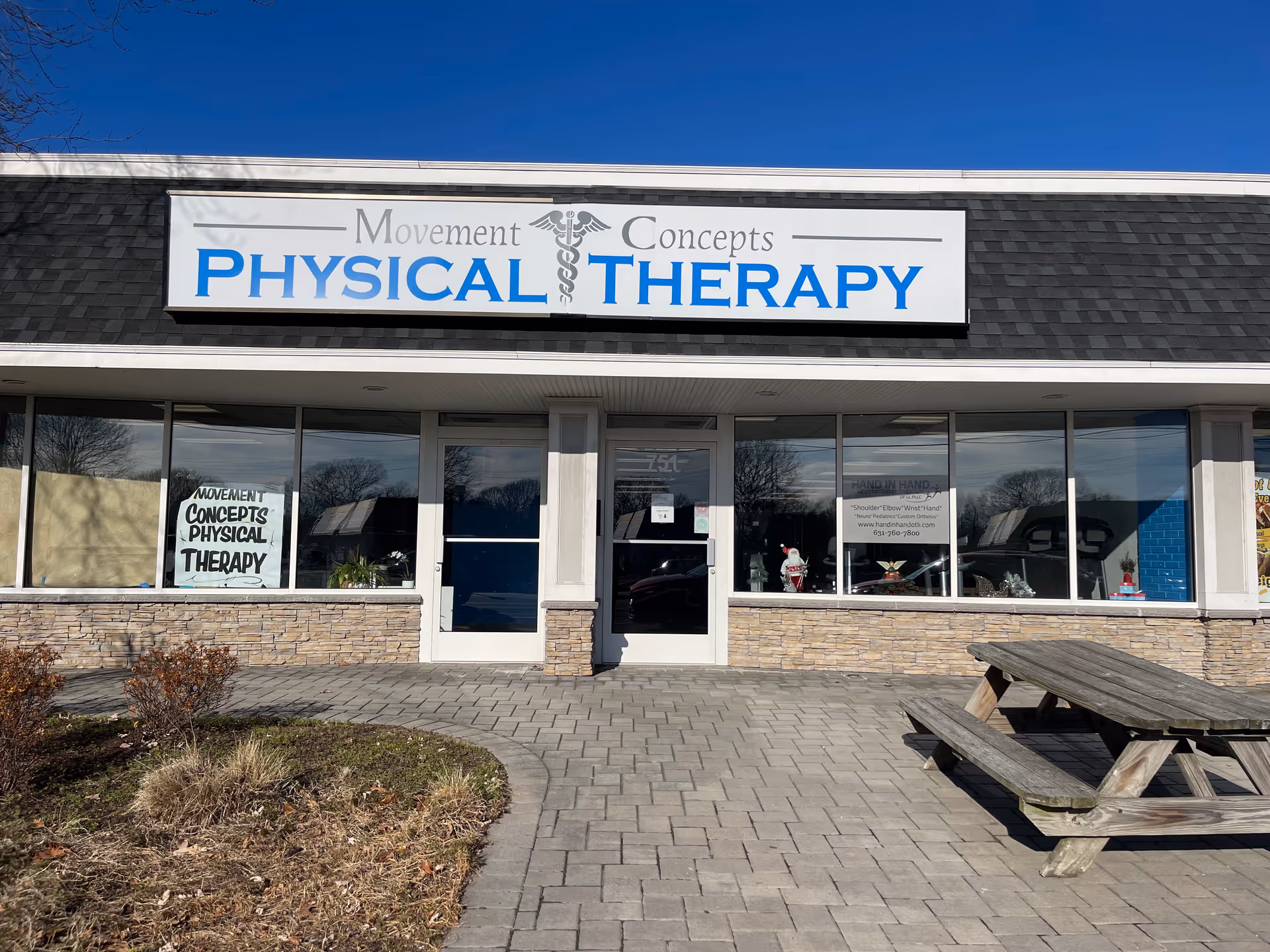 Exterior of Movement Concepts Physical Therapy storefront with large sign, windows, and a picnic table.