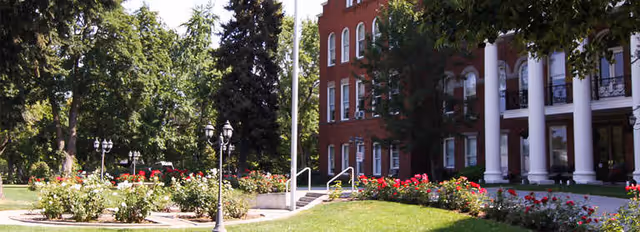 Front entrance of a red-brick building with white columns, surrounded by rose gardens, lawn and lampposts.