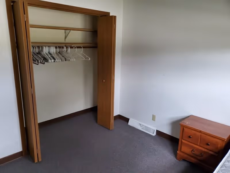 A small room with an open wooden closet containing several empty hangers. The room has plain white walls, a gray carpeted floor, a wooden nightstand with two drawers, and a white floor vent near the baseboard.
