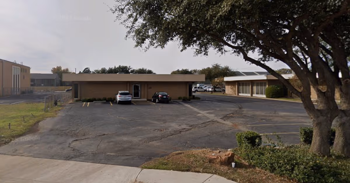 Two connected single-story buildings with a parking lot and a large tree.
