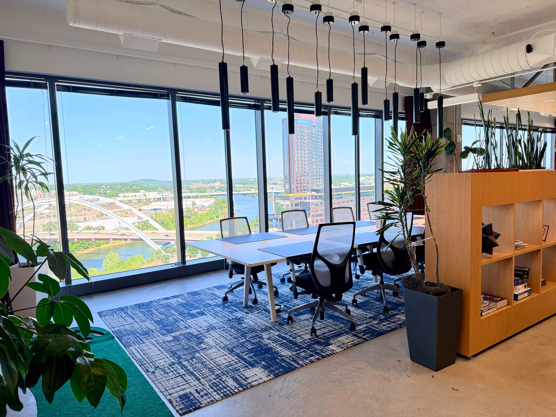 Bright office conference area with glass windows, desks, chairs, and tall plants.