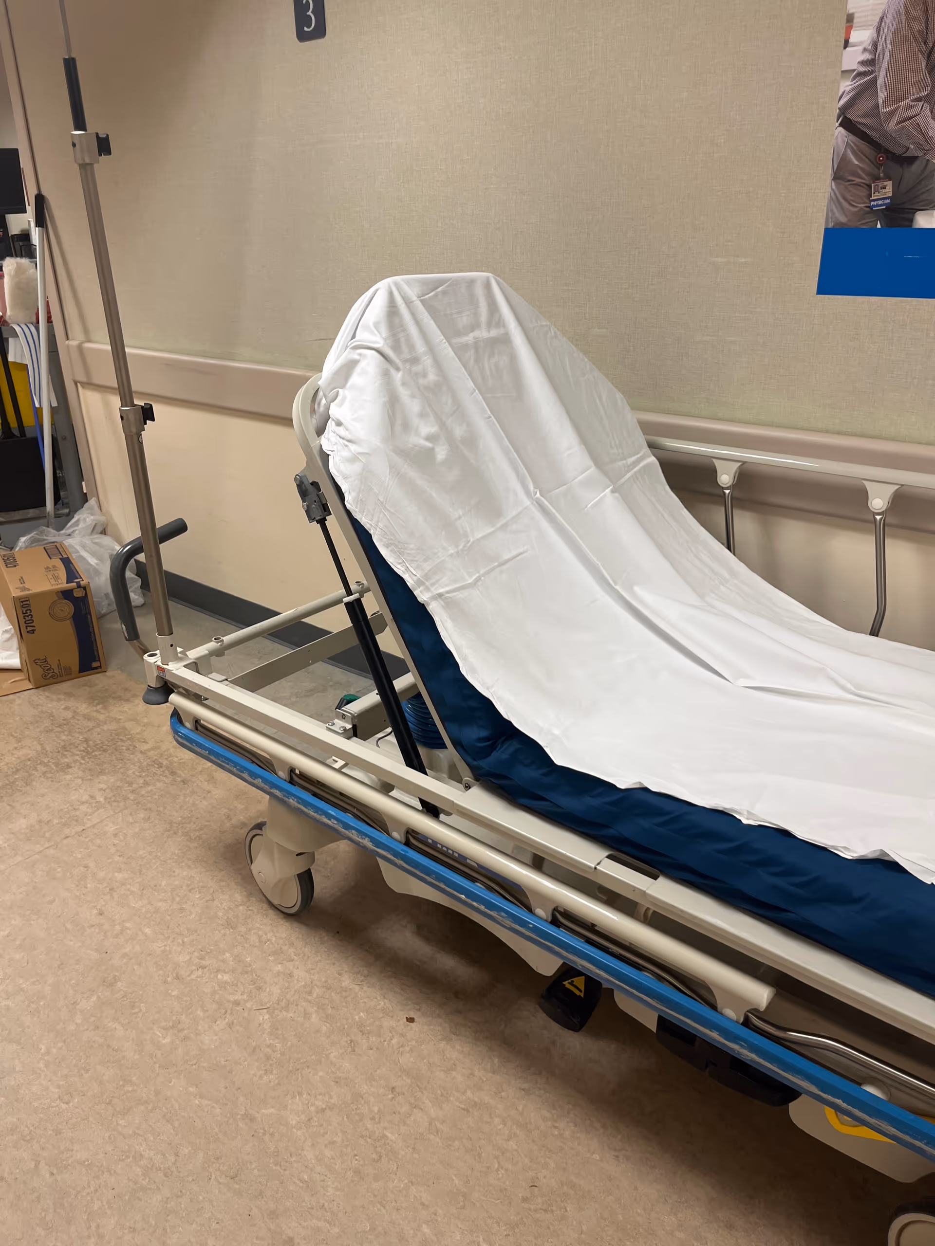 Hospital stretcher covered with sheet sits in a hallway near patient rooms.