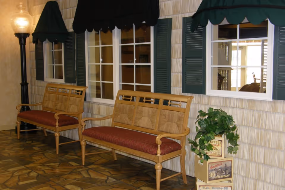 Indoor seating area with two wooden benches featuring red cushions placed against a wall with three windows that have green shutters and dark green awnings. A tall black lamp stands on the left side, and a decorative plant in a pot is on the right side near the benches. The floor has a stone tile pattern.