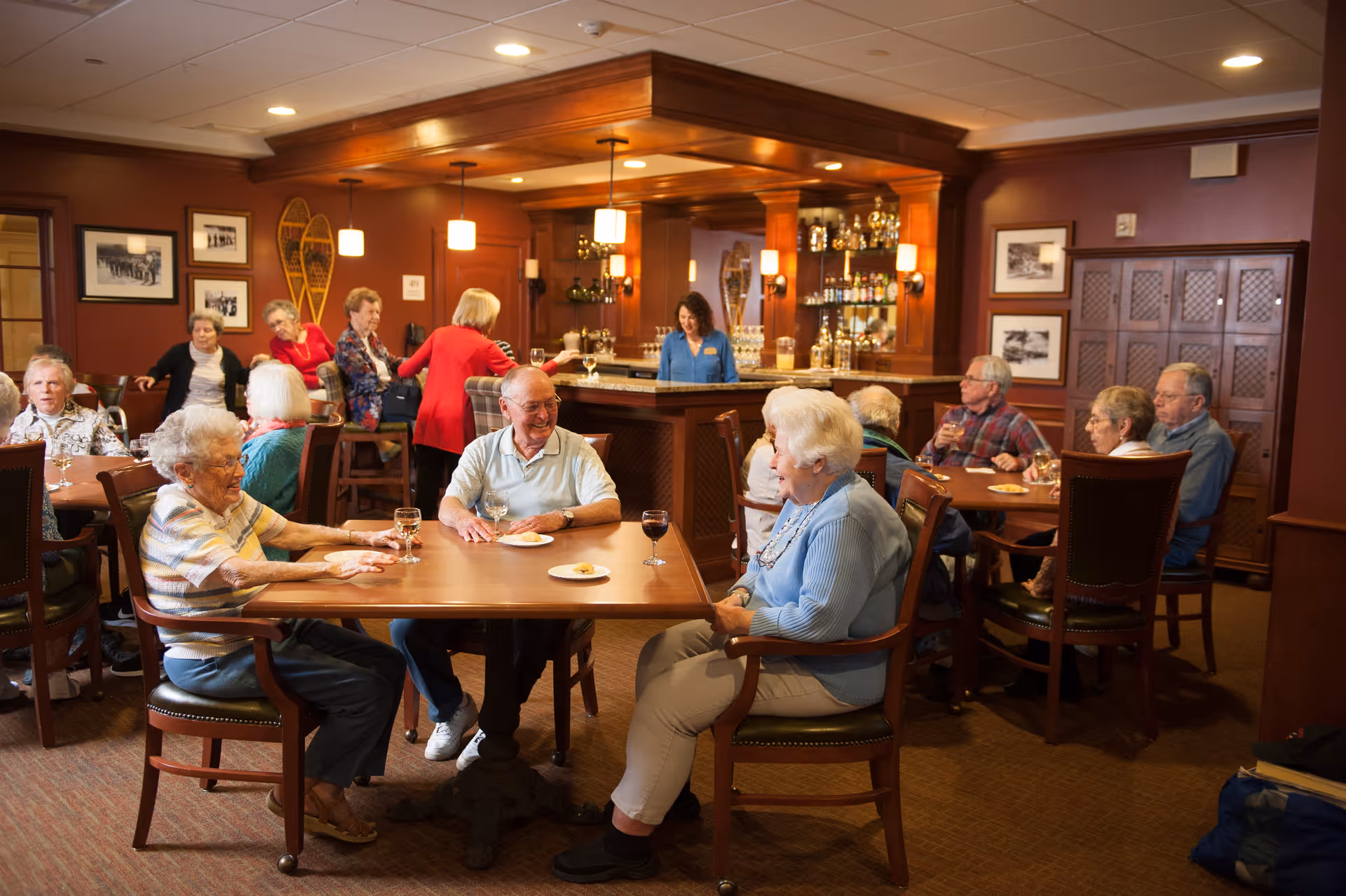 A group of elderly people sitting and socializing at tables in a warmly lit room with wooden furniture and a bar in the background where a staff member is standing. The room has framed pictures on the walls and a cozy, inviting atmosphere.