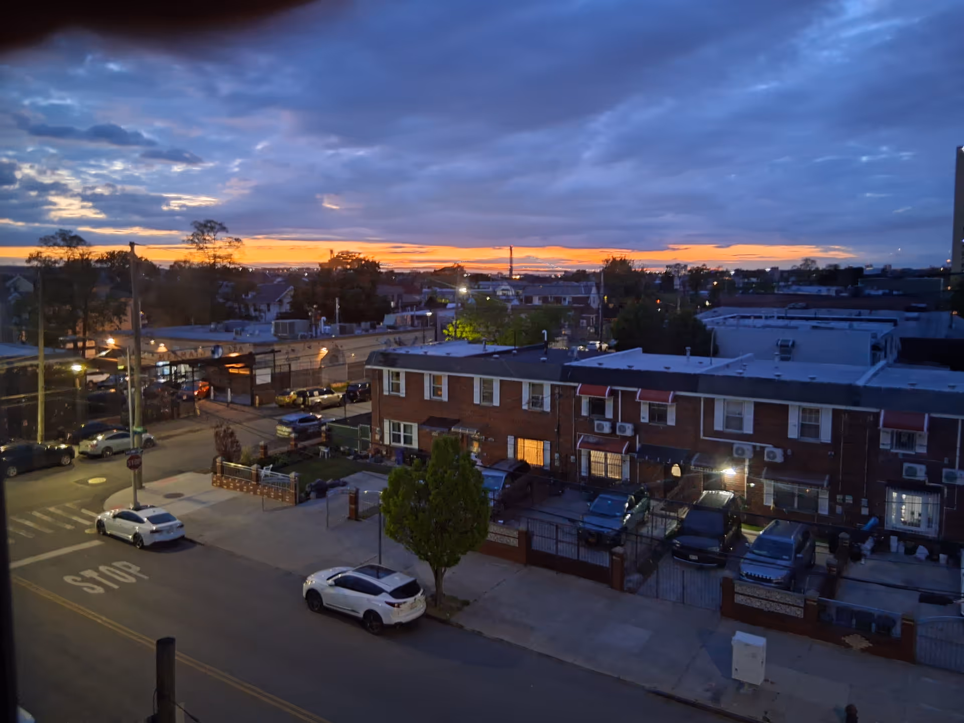Sunset over a row of brick townhouses with parked cars along a city street.