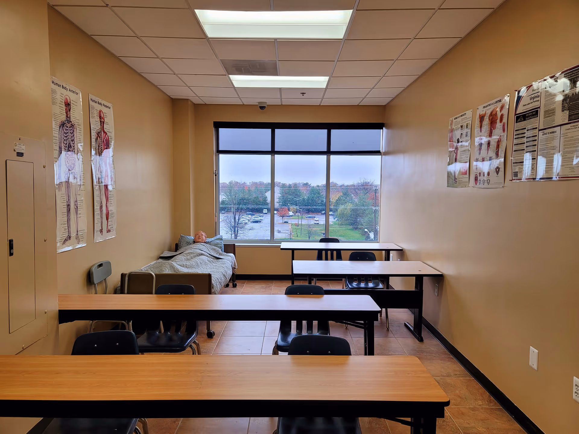 A classroom-style room in a senior facility with desks, chairs, posters, and a patient in bed.
