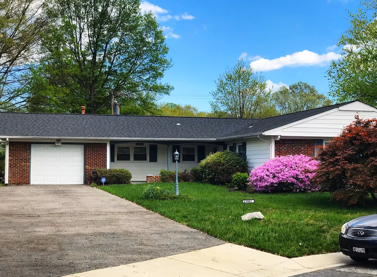Single-story suburban building with a front lawn, driveway, and flowering shrubs under a blue sky.
