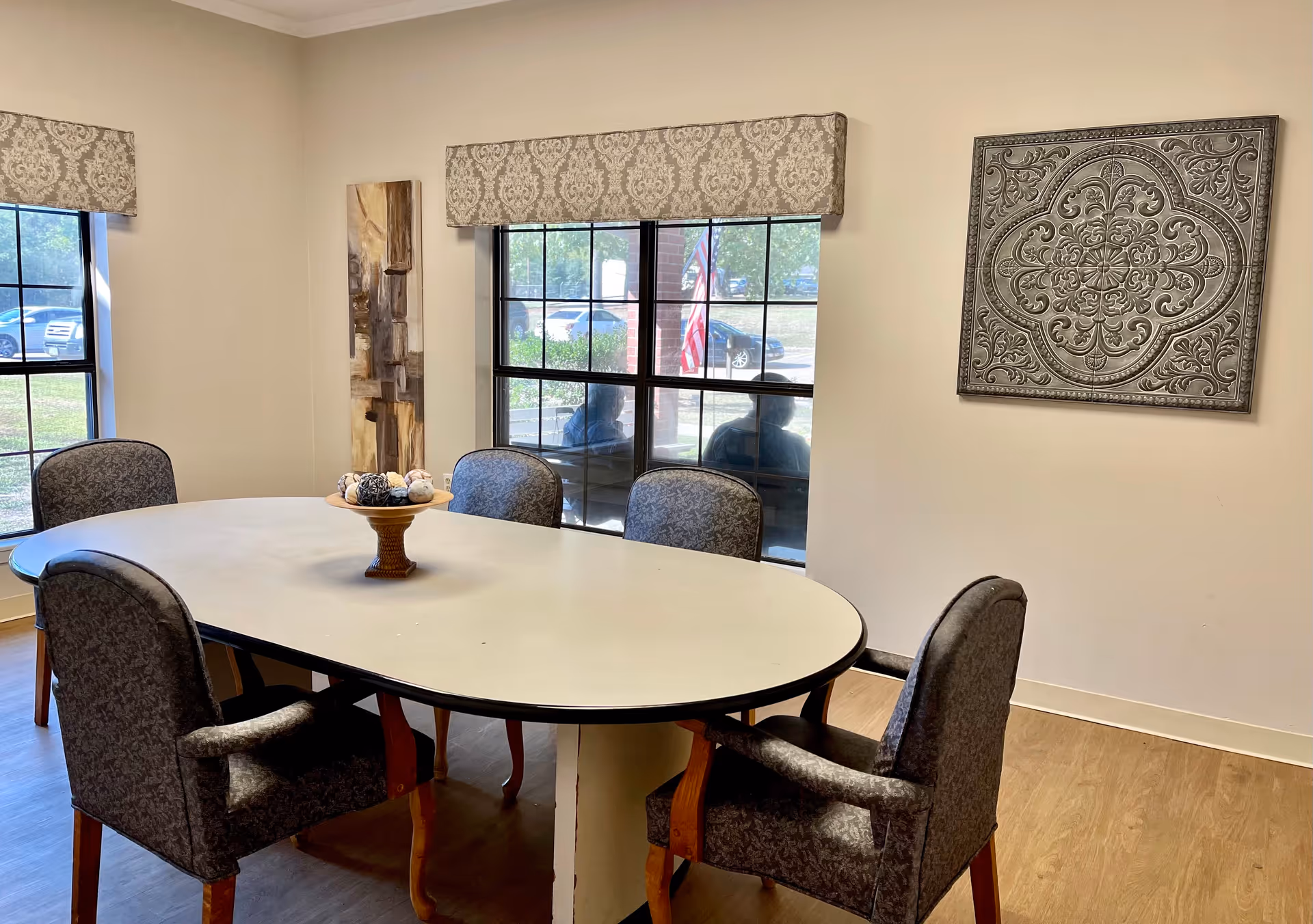 A small dining area with an oval white table surrounded by six upholstered chairs with wooden legs. The room has two windows with patterned valances, and outside the windows, cars and an American flag are visible. On the wall, there is a decorative metal wall art piece and a tall abstract painting. The floor is wood, and the room is well-lit with natural light.