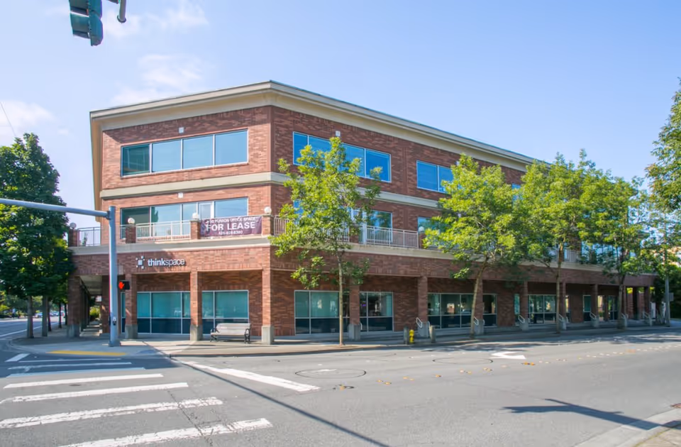 Three-story brick building on a street corner with trees, sidewalks, and storefront windows.