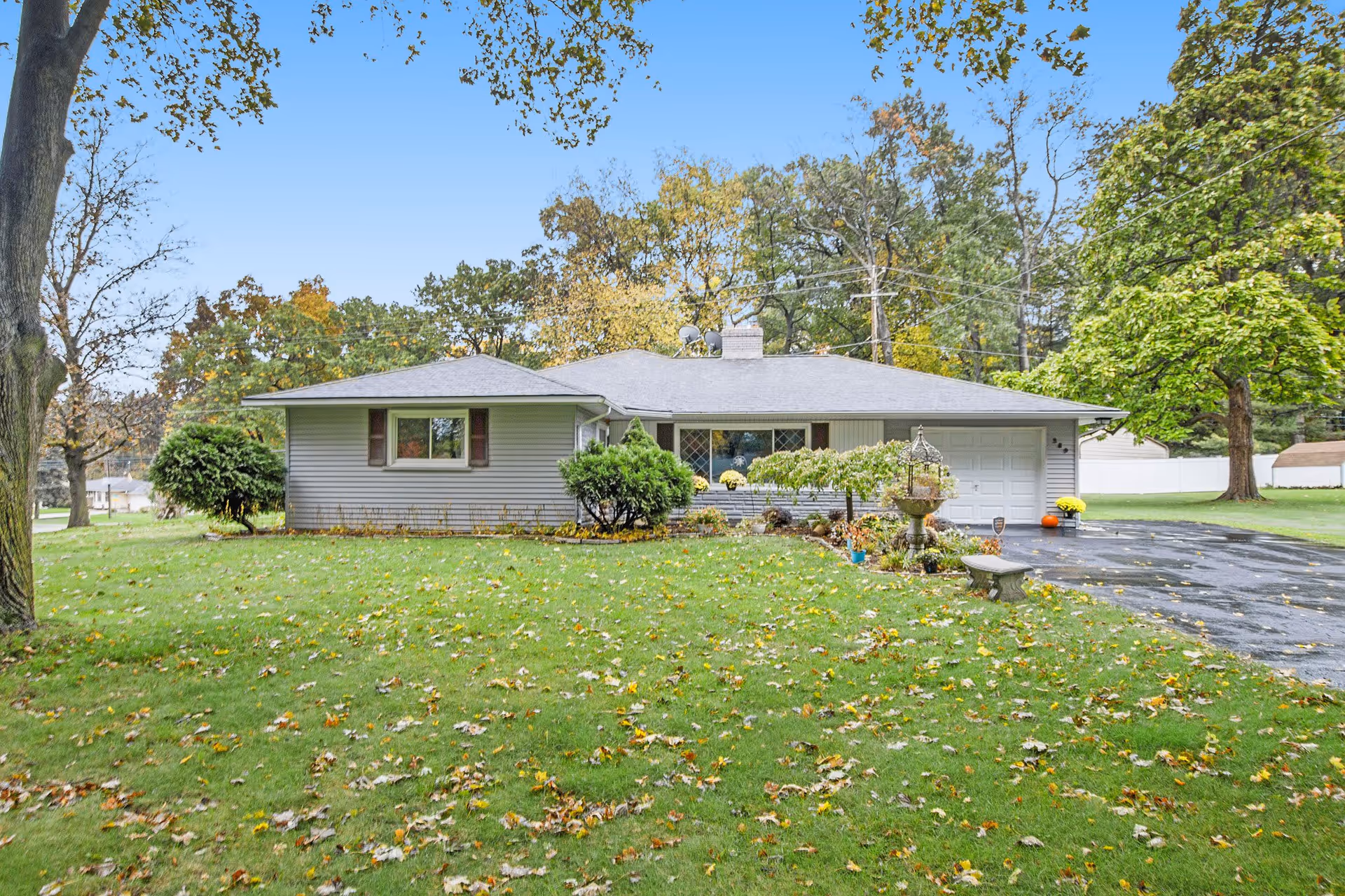 Single-story house with a gray roof and light-colored siding, surrounded by a green lawn with scattered fallen leaves. There are bushes and small trees near the house, a driveway on the right side, and a large tree on the right edge of the image. The sky is clear and blue.
