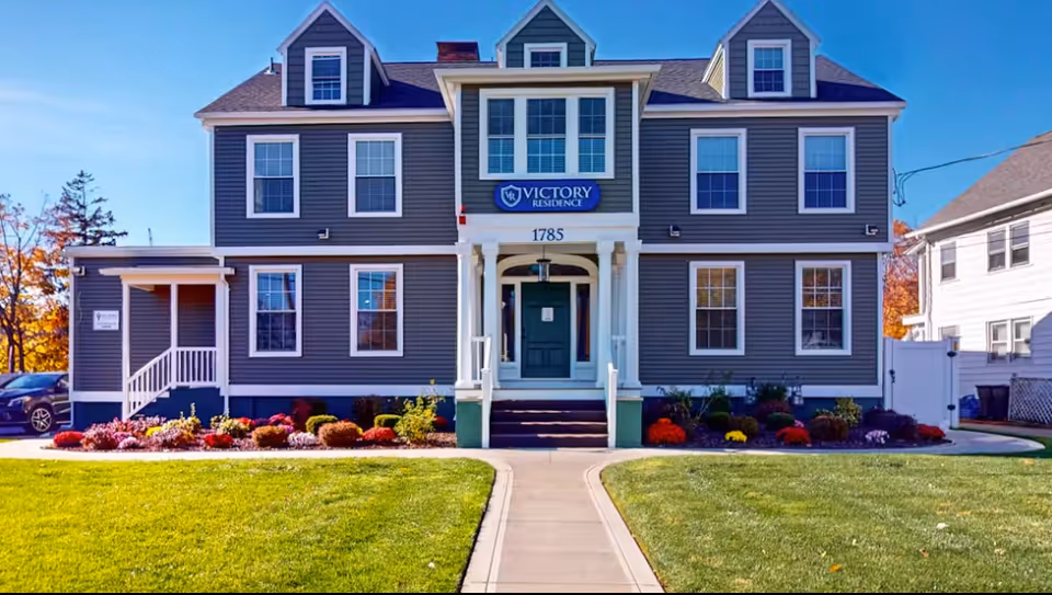 The front facade of a gray, multi-story senior residence with steps, a walkway, and landscaped lawn.