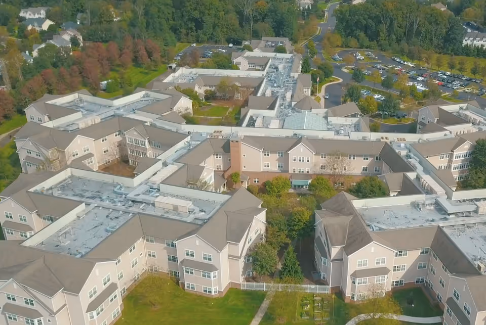 Aerial view of a large senior living facility complex surrounded by trees and parking lots, showing multiple interconnected beige buildings with gray roofs and green lawns.