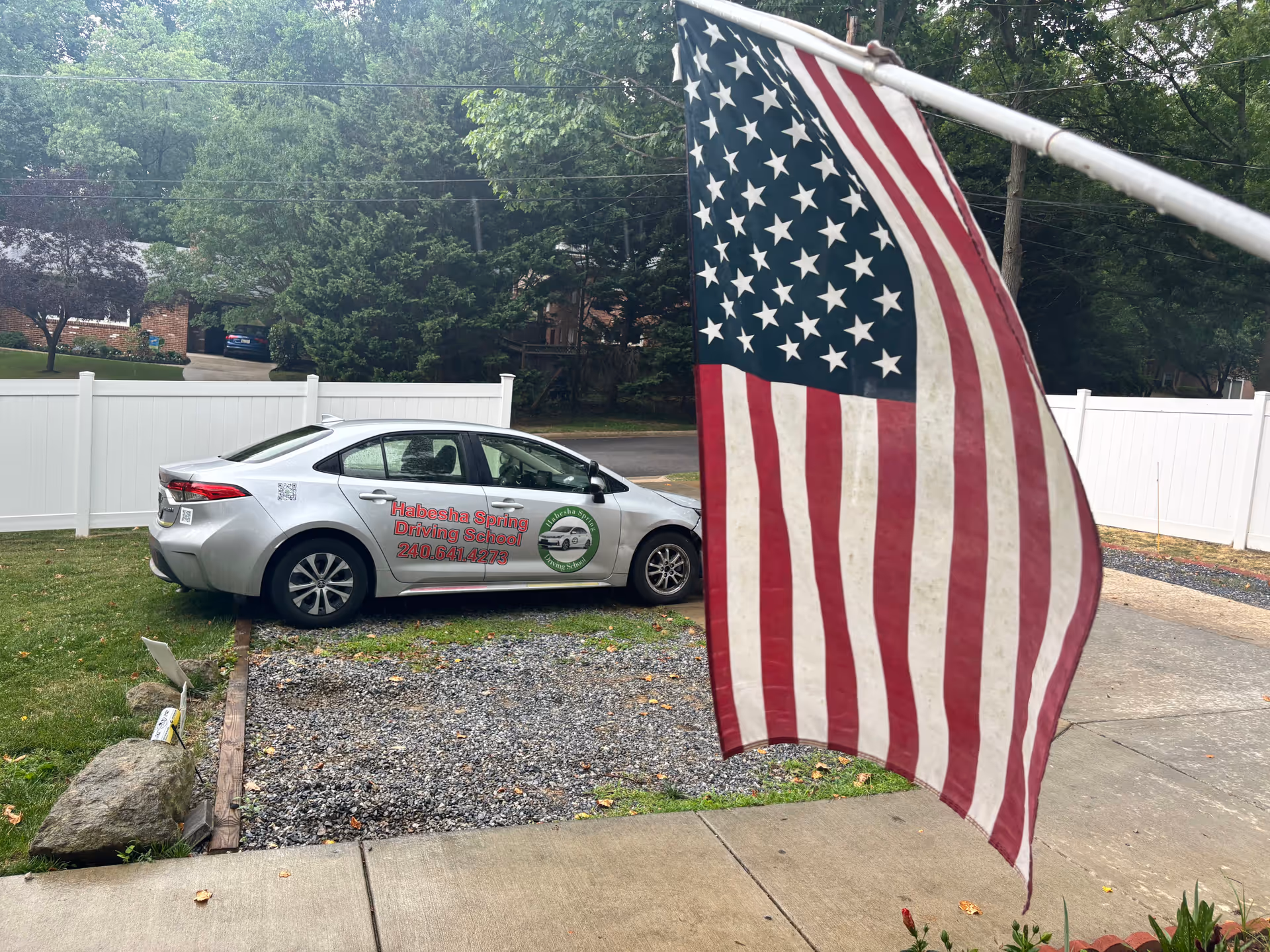 Silver sedan with driving-school decals parked beside a white fence, near an American flag.