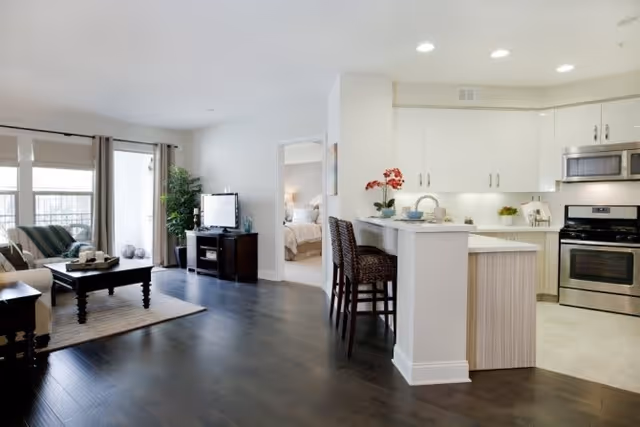 Open-plan interior showing a living room with sofa and TV and an adjacent kitchen with an island and bar stools.