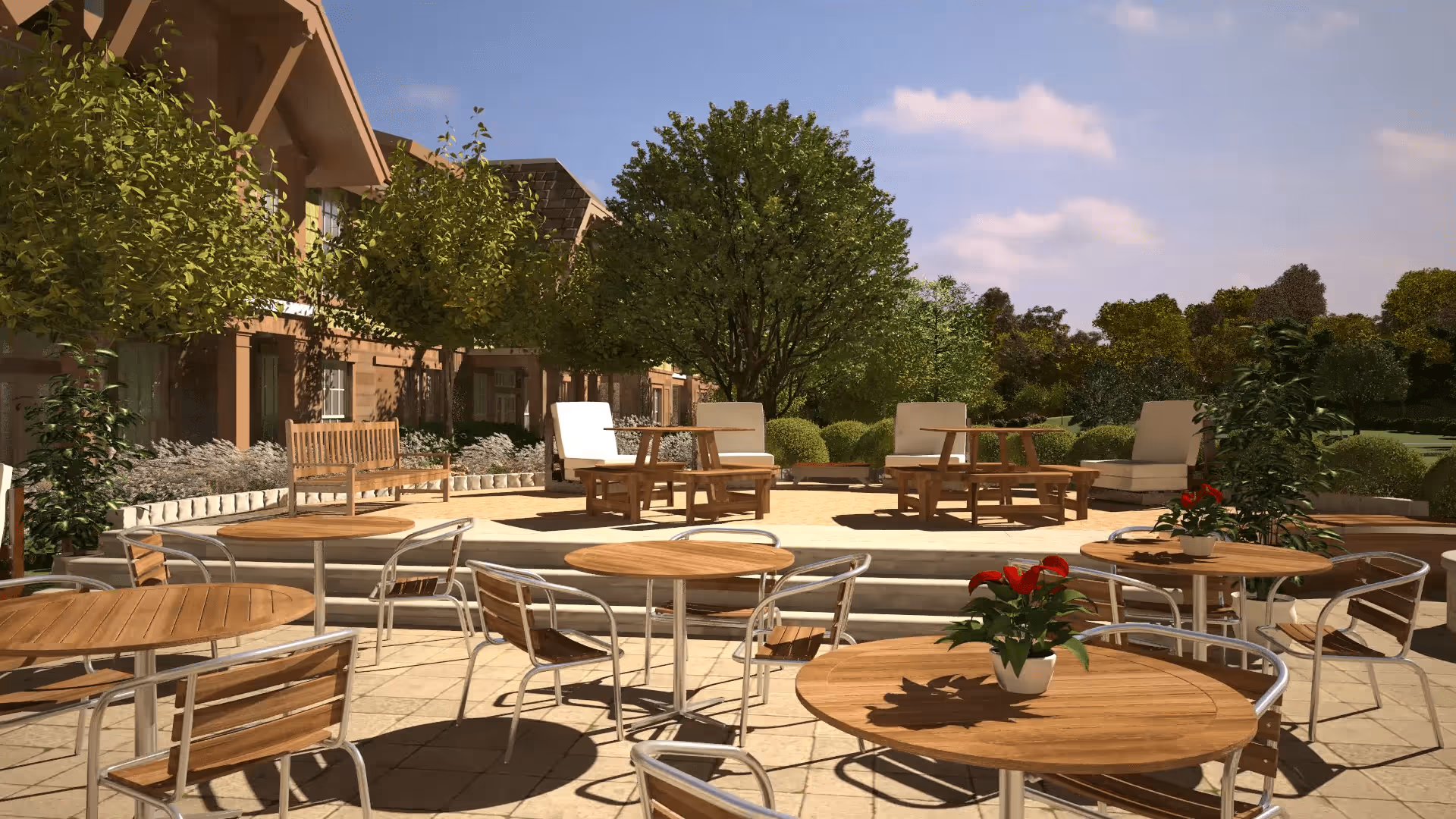 Outdoor patio area with round wooden tables and metal chairs, some tables decorated with small potted plants with red flowers. In the background, there are cushioned lounge chairs and wooden benches surrounded by green trees and bushes under a partly cloudy sky.