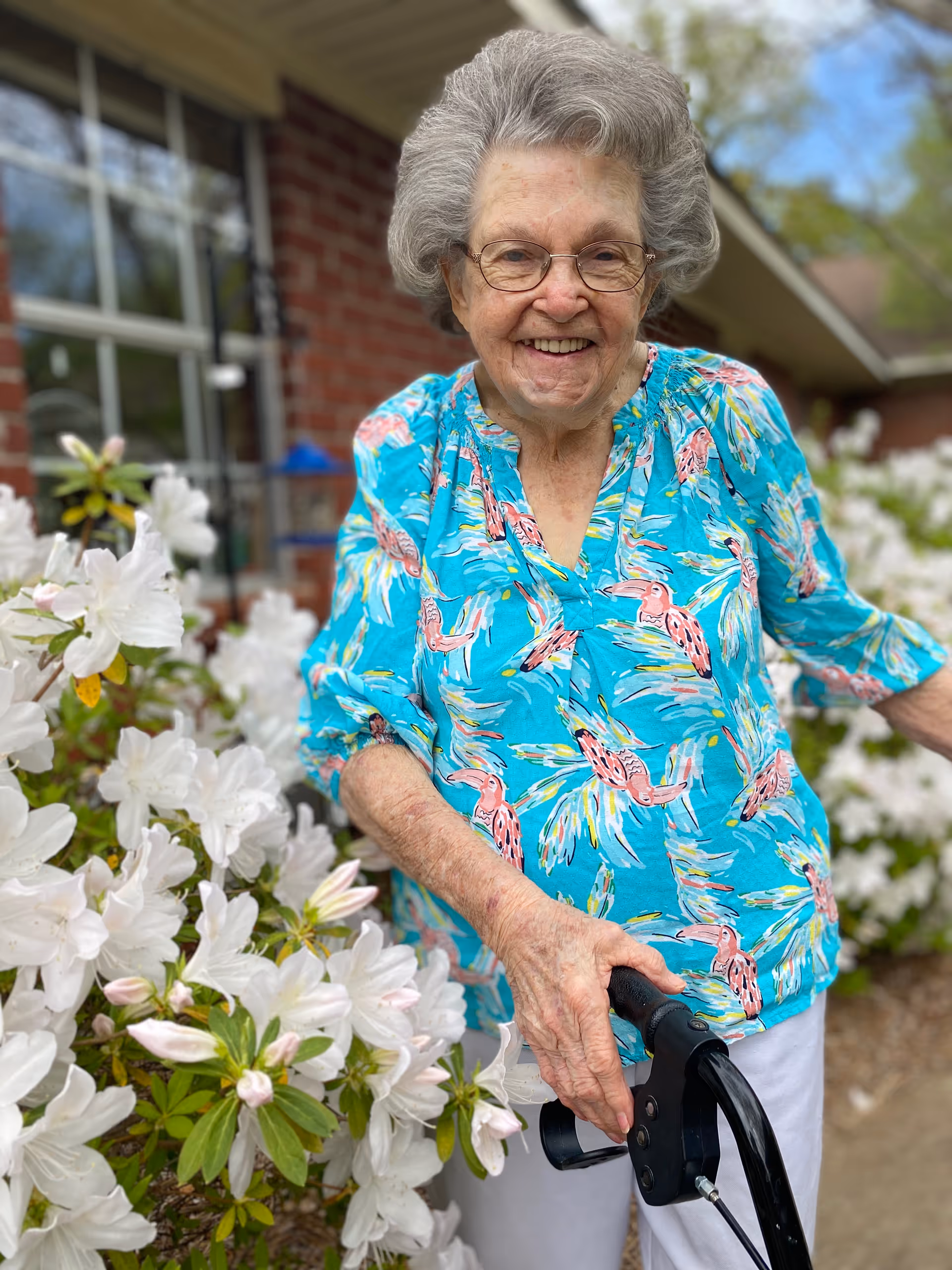 An elderly woman with gray hair and glasses is smiling while standing outdoors next to white flowering bushes. She is wearing a bright blue blouse with a colorful bird pattern and holding a black walker. A brick building and window are visible in the background.