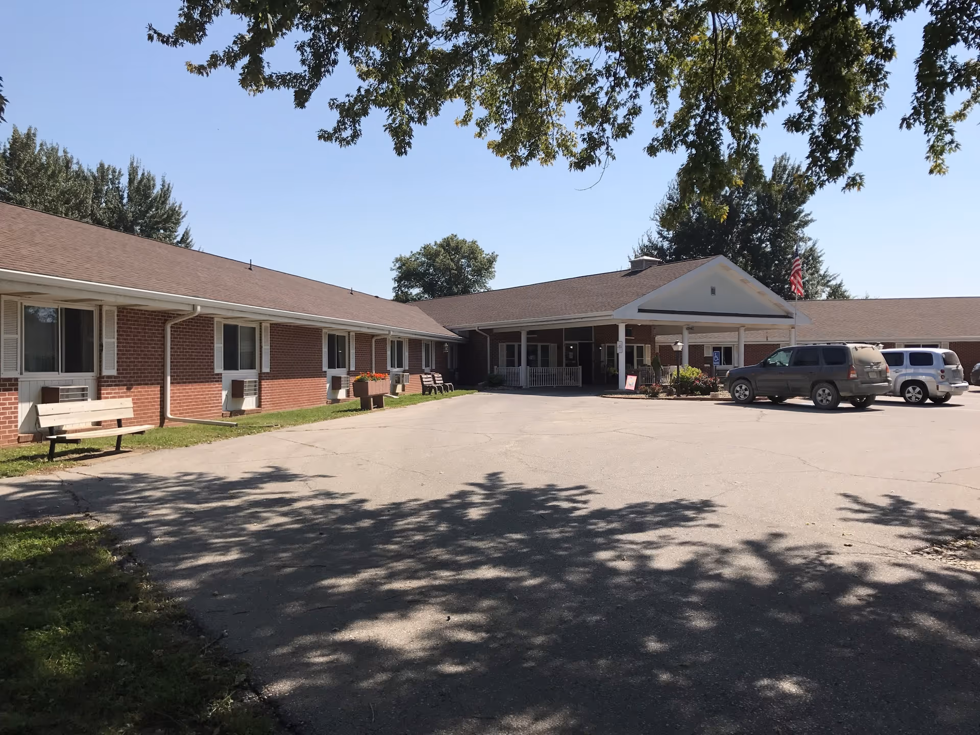 Exterior view of Denison Health Care Center showing a single-story brick building with a covered entrance, several windows, benches along the side, a few parked vehicles, and trees casting shadows on the pavement.