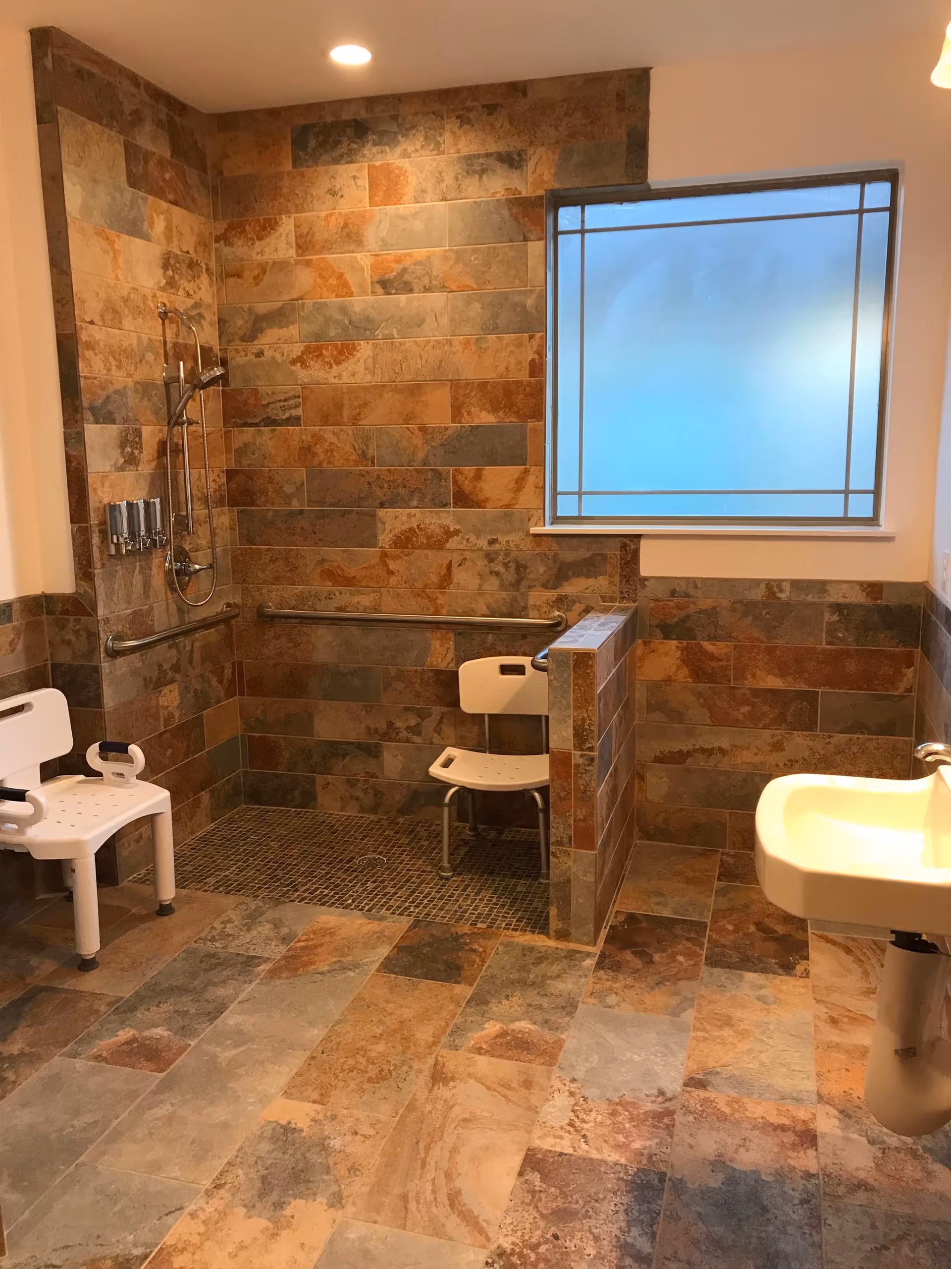 Tiled accessible bathroom featuring a roll-in shower with grab bars and shower chairs, a frosted window, and a wall-mounted sink.