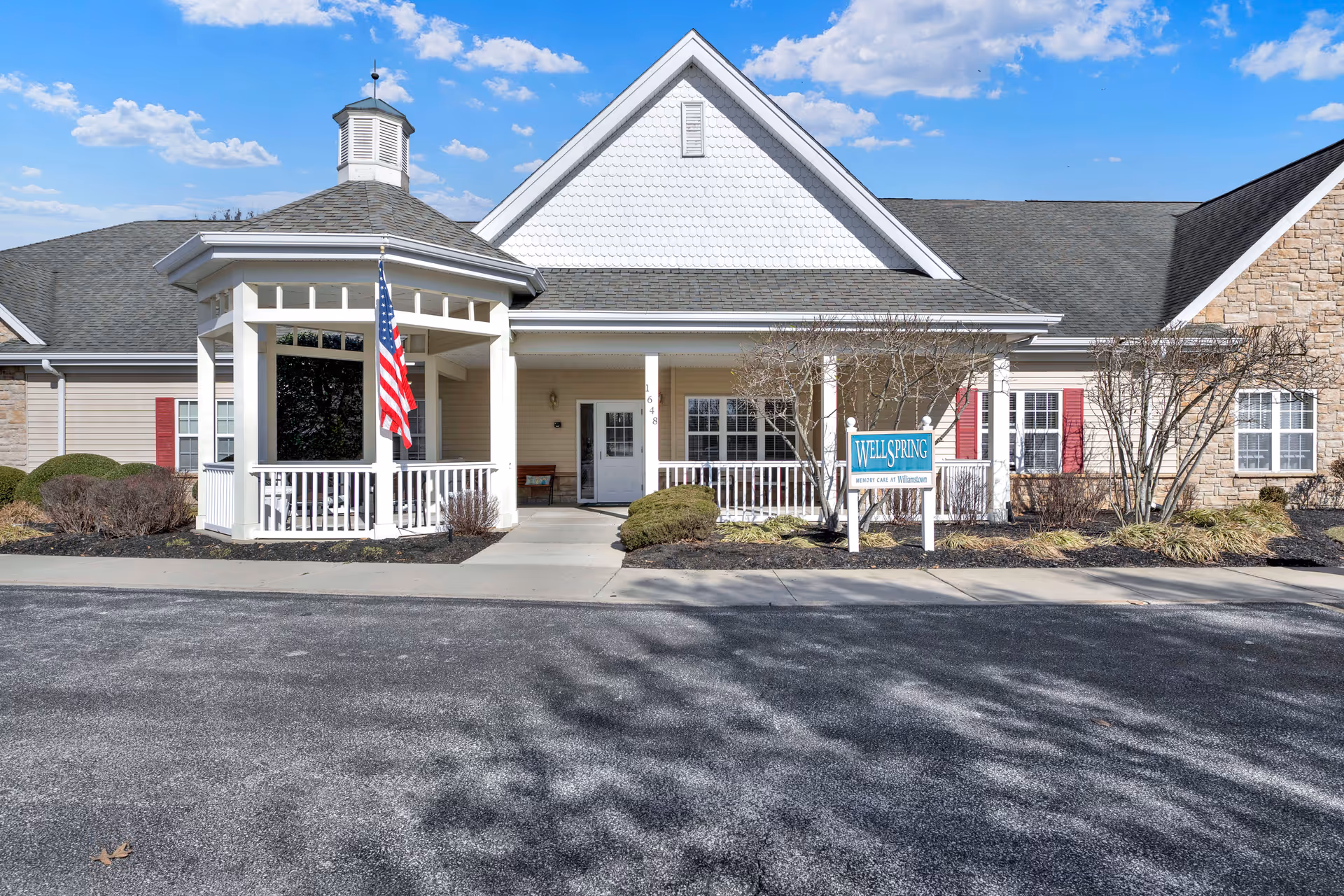 Front exterior view of a senior living facility building with a white gazebo displaying an American flag, a porch with white railings, and a sign that reads 'WELLSPRING Memory Care at Williamstown'. The building has beige siding, stone accents, and red shutters under a blue sky with some clouds.