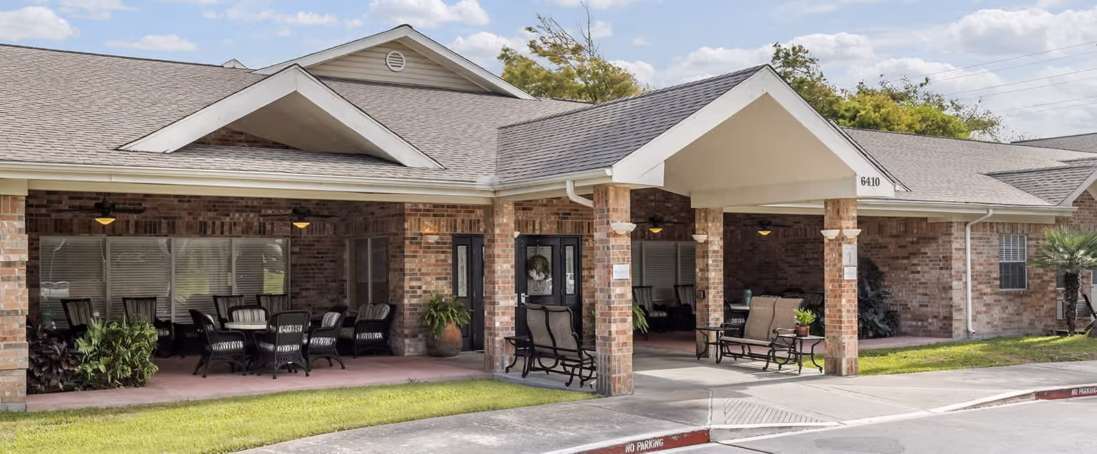 Exterior view of a single-story brick building with a covered entrance supported by brick columns. The entrance area has outdoor seating with chairs and tables. The building has a gabled roof and is surrounded by a well-maintained lawn and some plants.