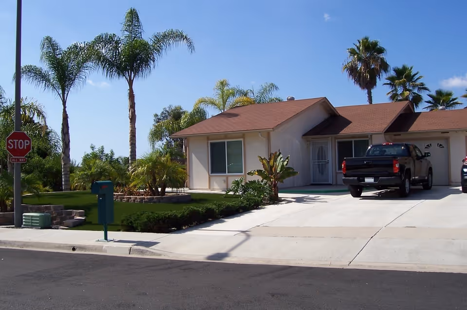 Exterior view of a single-story residential building with a brown roof and beige walls, a driveway with a black pickup truck parked, palm trees and other greenery around the property, and a stop sign at the street corner.