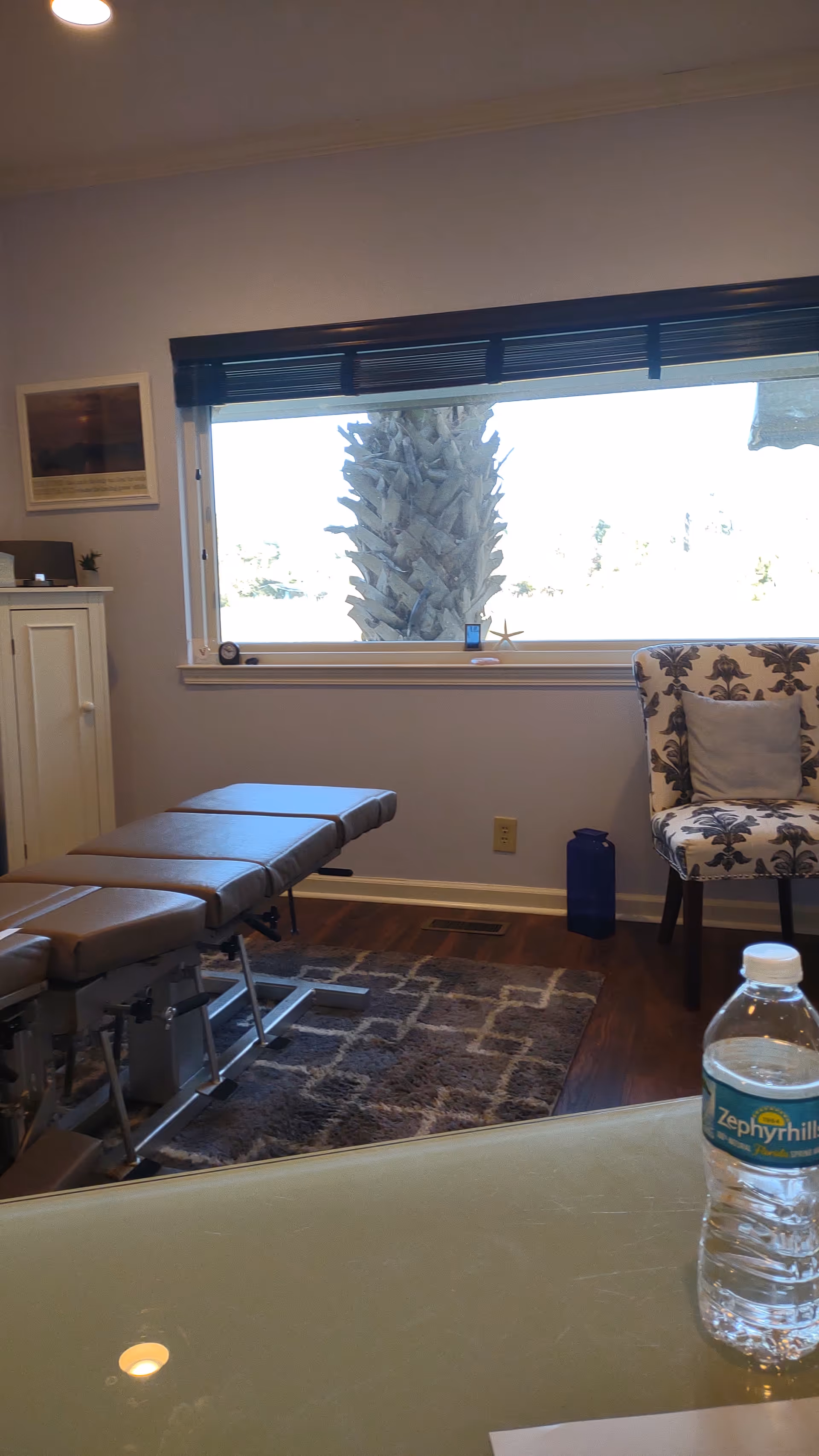 Chiropractic treatment room with adjustable table, window view, patterned chair, and water bottle.