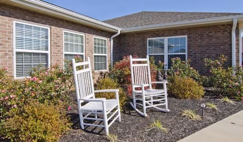 Two white rocking chairs sit on mulch in front of a brick building with shrubs.