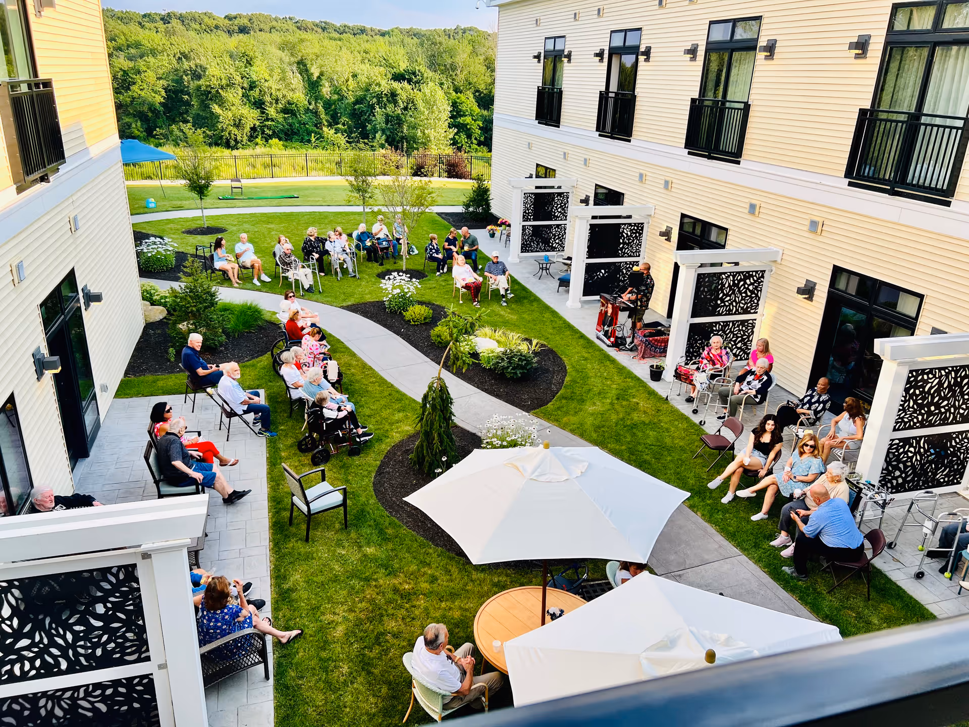 Outdoor courtyard area of Briarcliffe Preserve Assisted and Independent Living with many elderly residents sitting on chairs and benches arranged along a curved pathway surrounded by grass, plants, and small trees. The building with balconies and decorative privacy panels is visible on both sides, and there are large white umbrellas providing shade over tables.
