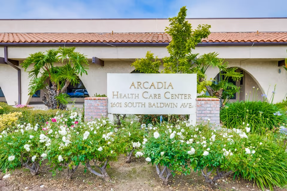 Front exterior view of Arcadia Health Care Center with a sign displaying the facility name and address, surrounded by green plants and white flowers, with a building featuring arched windows and a tiled roof in the background.