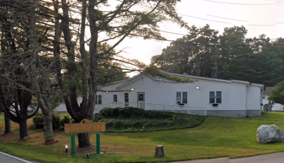 A single-story white senior living facility with trees, a lawn, and a sign.