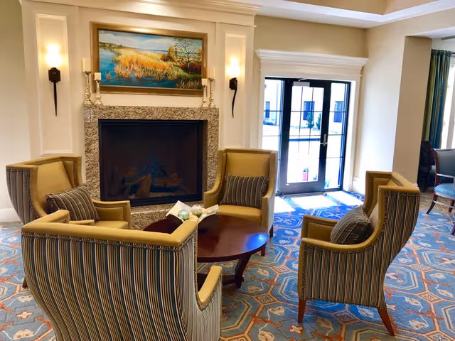 A cozy seating area in a senior living facility with five striped armchairs arranged around a wooden coffee table. The room features a fireplace with a granite surround, a landscape painting above it, wall sconces on either side, and a glass door leading outside. The floor is covered with a patterned carpet.
