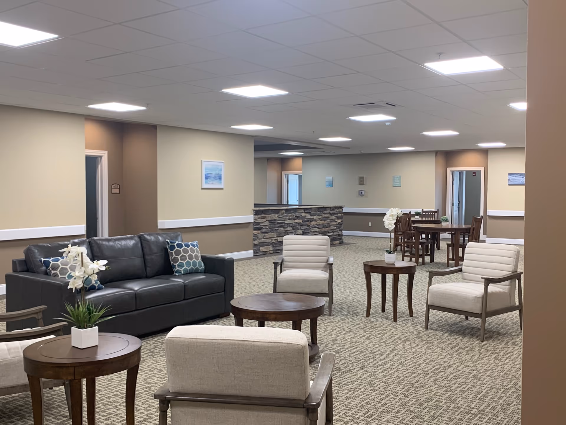 Spacious senior living lounge with sofas, chairs, tables, and a stone-front reception area.