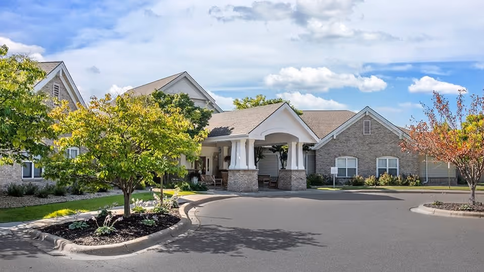 Exterior view of a senior living facility with a covered entrance, surrounded by trees and landscaping under a partly cloudy sky.
