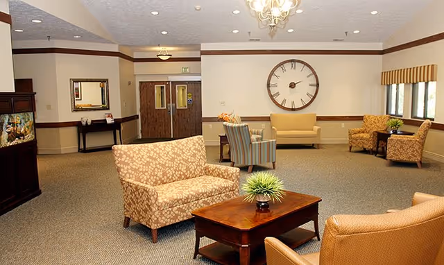 Neutral-toned senior living facility lounge with sofas, chairs, coffee table, and a large wall clock.