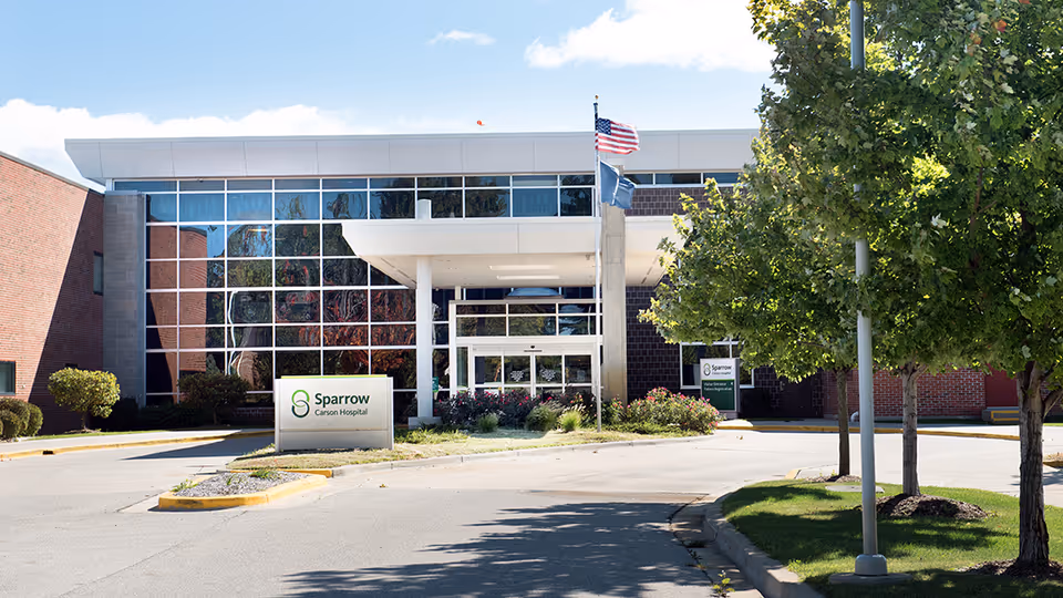 Modern medical facility entrance with glass facade, flags, trees, and a Sparrow Carson Hospital sign