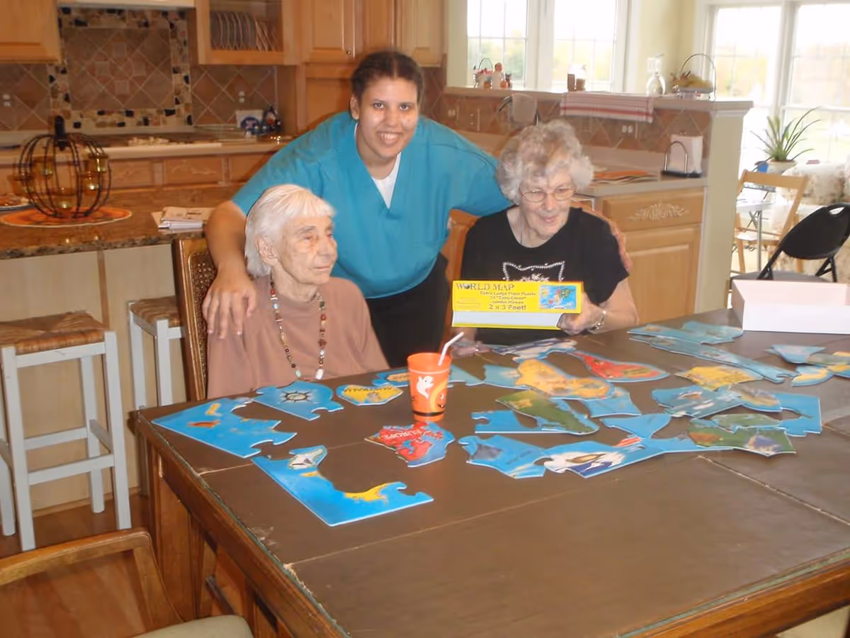 Two elderly women sitting at a table working on a large world map puzzle with a caregiver standing behind them smiling. The setting appears to be a kitchen or dining area with wooden cabinets and stools in the background.