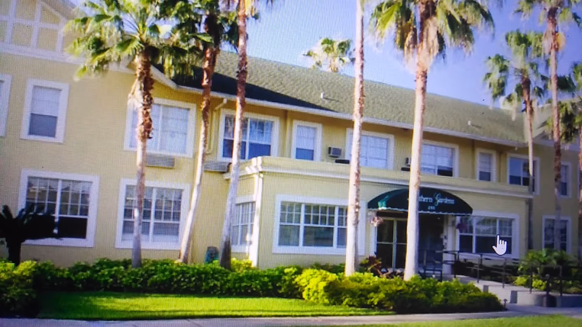 Two-story yellow senior living building with palm trees and a green awning entrance.
