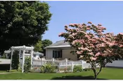 Exterior view of a single-story senior living facility with a porch, lawn, and pink flowering tree.
